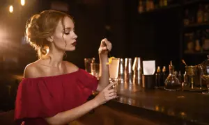 Elegancka kobieta w bordowej sukience, z idealnie dobranymi paznokciami, sączy drinka przy barze.
