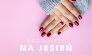 Dłonie z bordowymi, lśniącymi paznokciami na jesień, otulone szarym swetrem, na różowym tle.