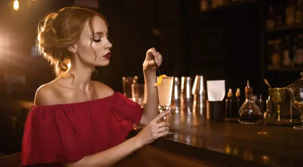 Elegancka kobieta w bordowej sukience, z idealnie dobranymi paznokciami, sączy drinka przy barze.