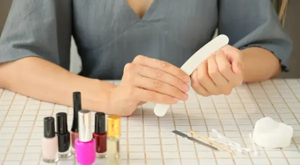 Kobieta wykonuje **manicure**, używając pilniczka do paznokci. Na stole stoją kolorowe lakiery i akcesoria do pielęgnacji.