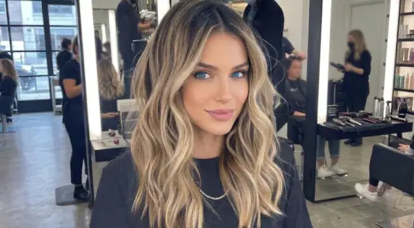 Kobieta z pięknymi, falowanymi blond włosami, z subtelnym, ciemnym odrostem przy blondzie. W tle salon fryzjerski.