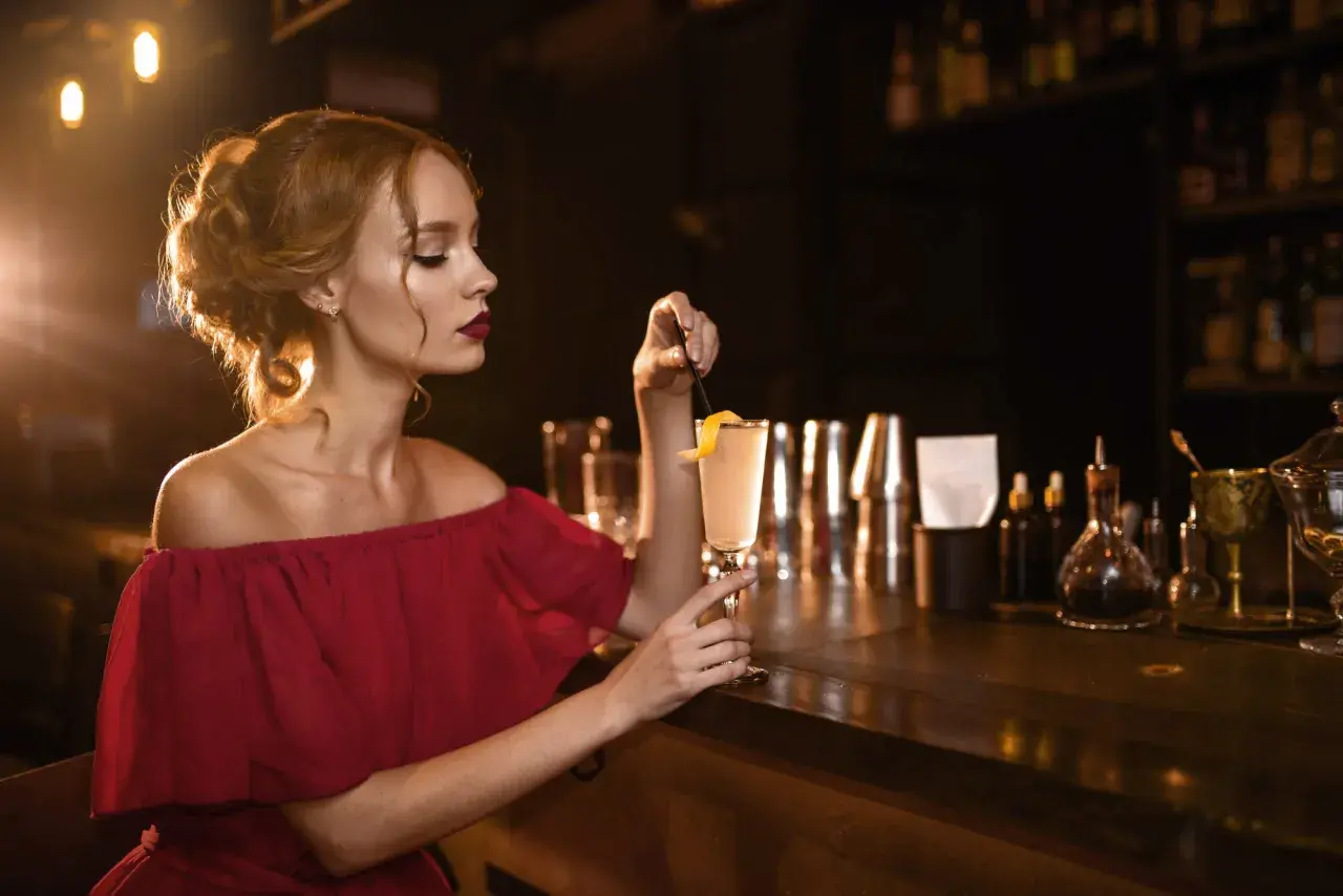 Elegancka kobieta w bordowej sukience, z idealnie dobranymi paznokciami, sączy drinka przy barze.
