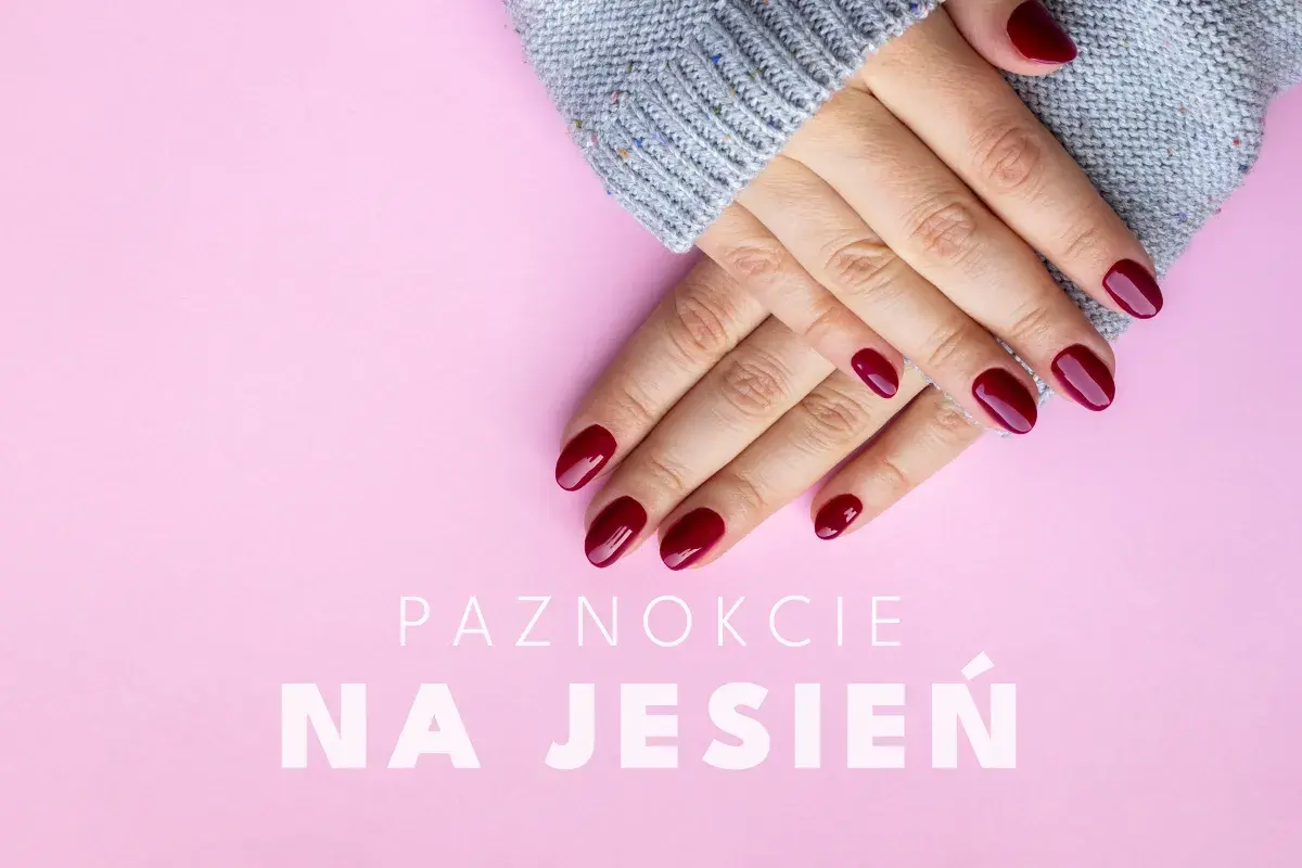 Dłonie z bordowymi, lśniącymi paznokciami na jesień, otulone szarym swetrem, na różowym tle.