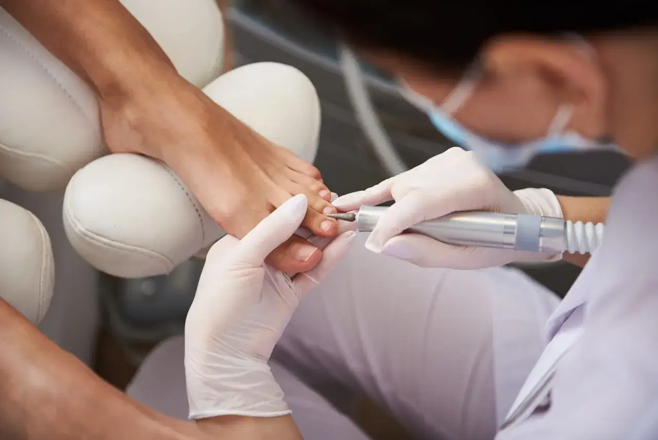 Profesjonalny pedicure frezarkowy w trakcie. Dłoń w rękawiczce z frezarką pracuje przy paznokciu stopy.