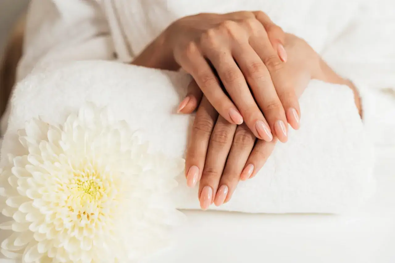 Delikatne dłonie z paznokciami w cielistym kolorze, ułożone na białym ręczniku obok kwiatu. To efekt pięknego manicure, gdzie top coat zapewnia połysk i trwałość.