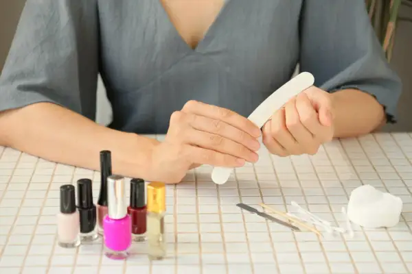 Kobieta wykonuje **manicure**, używając pilniczka do paznokci. Na stole stoją kolorowe lakiery i akcesoria do pielęgnacji.